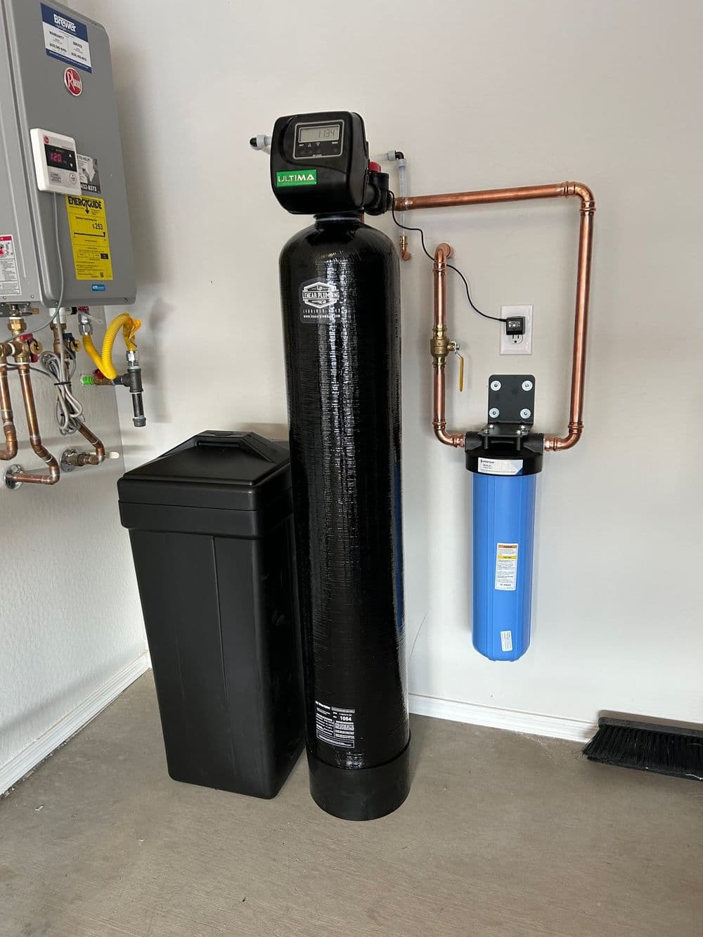 Water filtration system with a black tank, blue filter, and connected copper pipes in garage.